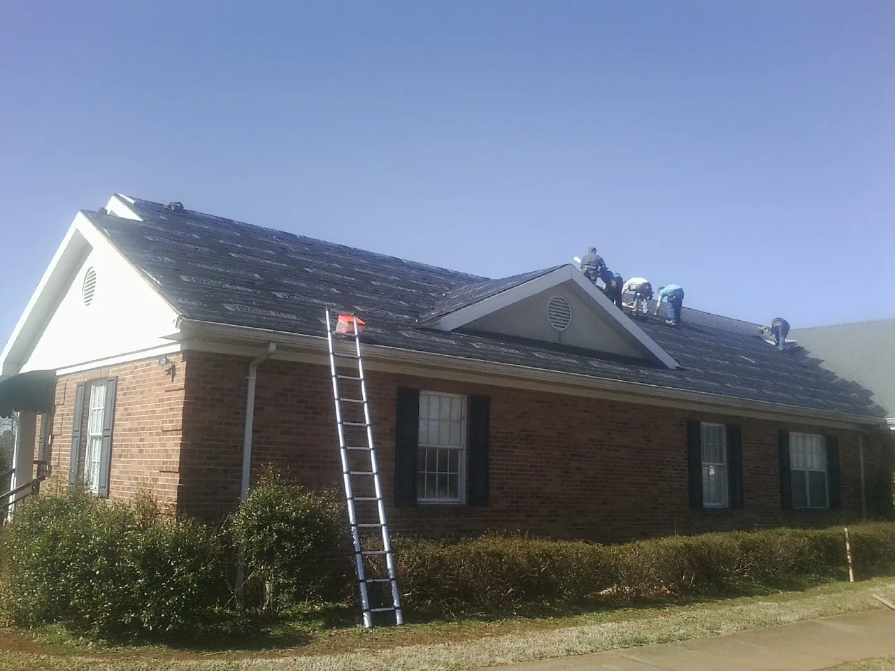 Crew tearing shingles off a brick ranch roof with a ladder set — Metal Roof Repair work by our crew in Otoe