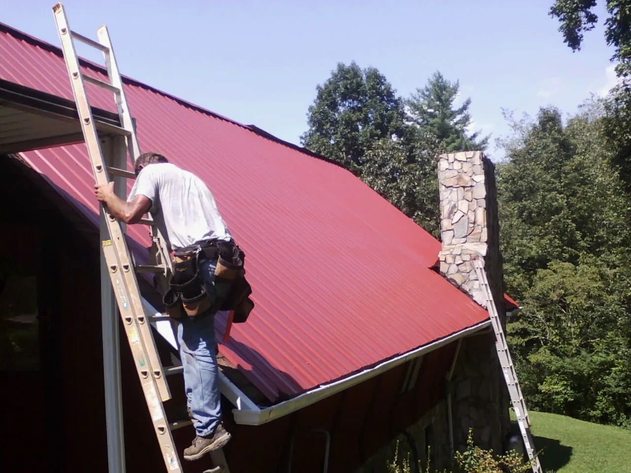 Roofer on a ladder beside a red metal roof and stone chimney — Soffit & Fascia Repair by Roofing Brothers in Otoe